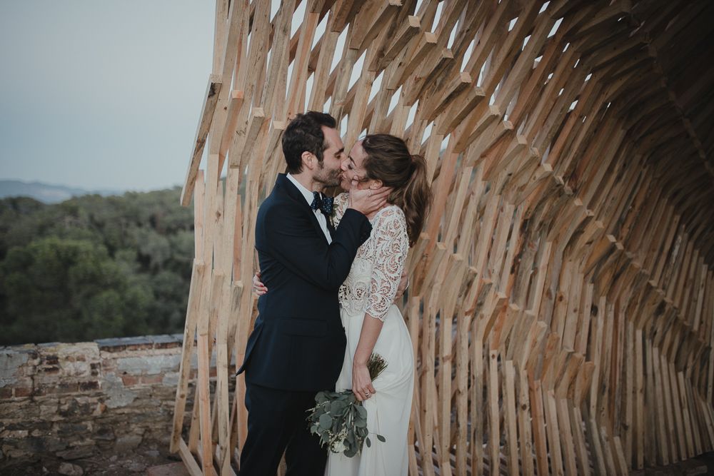 A romantic wedding moment captured under a curved wooden archway with a scenic backdrop of rolling hills.