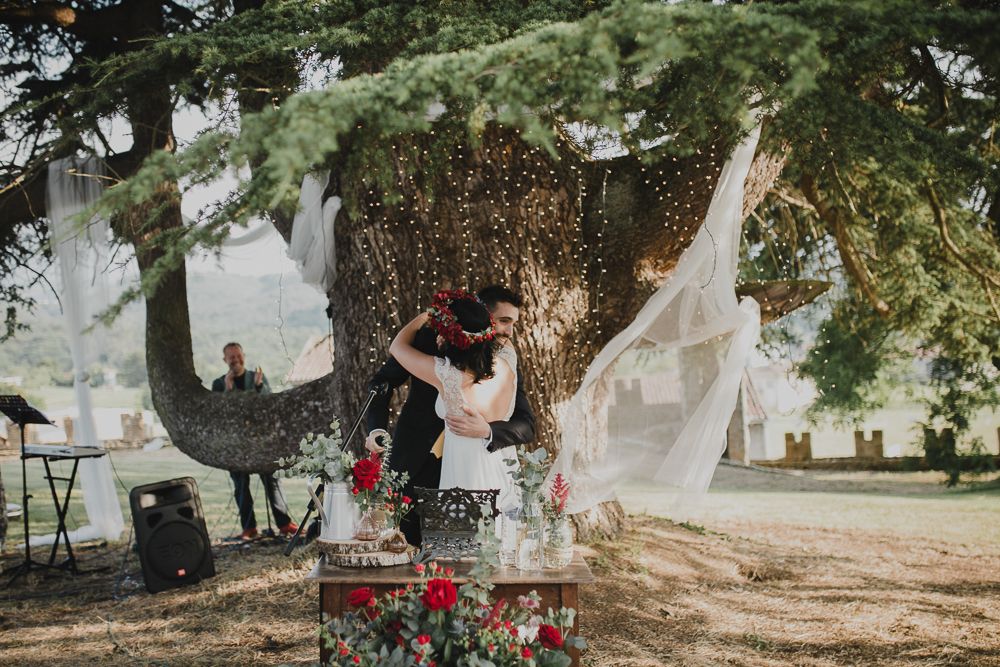 A romantic wedding scene under a large tree decorated with flowing white fabric and string lights.