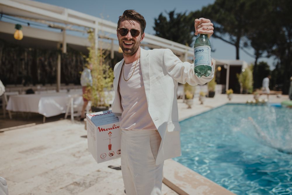 Person in white summer attire holding drinks by a swimming pool.