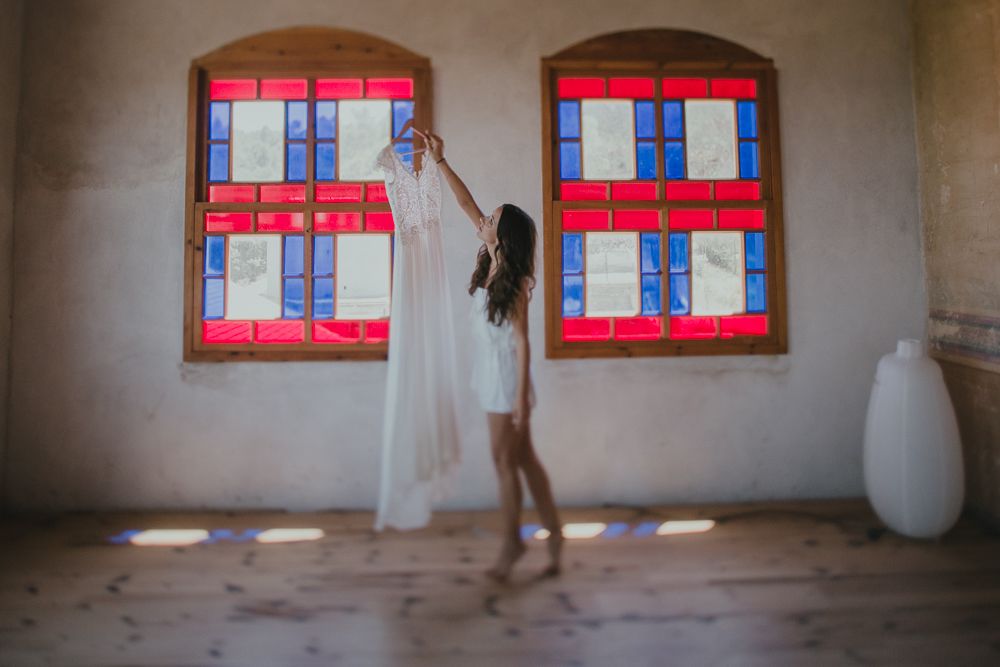 Figure in a white dress reaches up to vibrant red and blue shuttered windows in a rustic interior space.