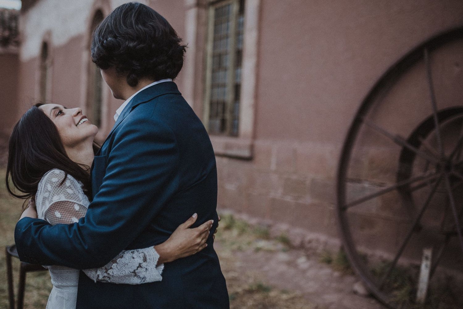An intimate embrace between a couple in formal attire against a rustic pink wall with an antique wheel.