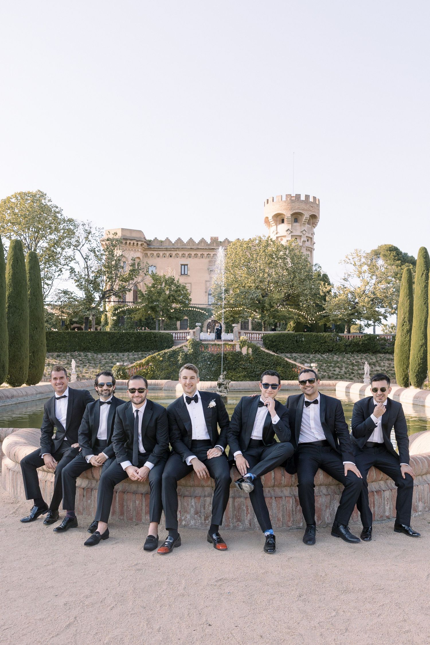 Wedding party members in formal attire sitting on stone wall with castle tower in background.