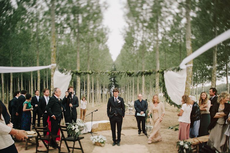 Wedding ceremony setup in a forest with rows of chairs and flowing white fabric decorations.