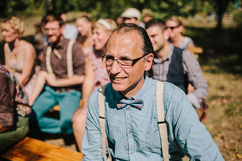 Guest in suspenders and bow tie smiles during outdoor wedding ceremony.