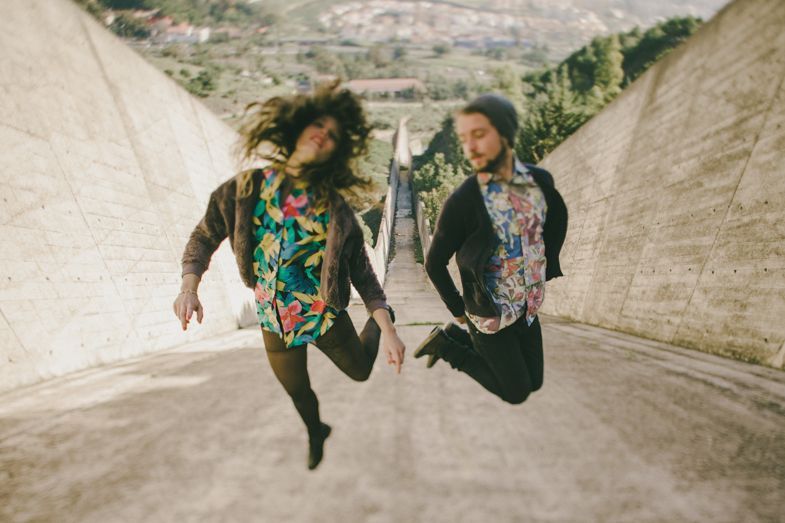 Two people jumping joyfully on concrete road wearing colorful patterned shirts.