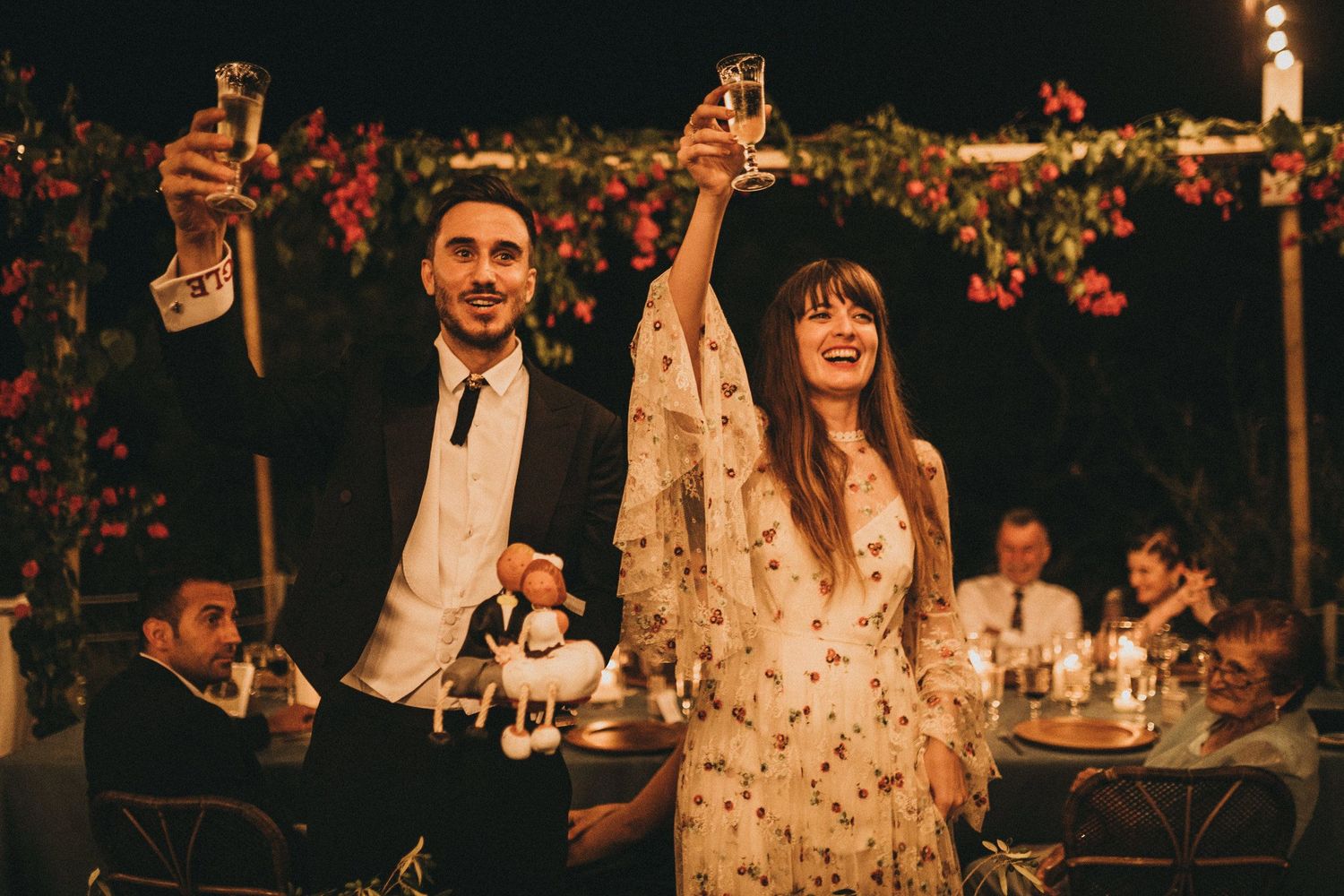 Celebratory toast at an outdoor evening reception under string lights and floral decorations.