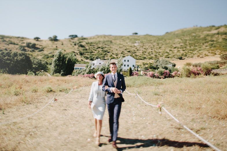 Wedding ceremony guests seated outdoors on a sunny hillside with mountains in the background.