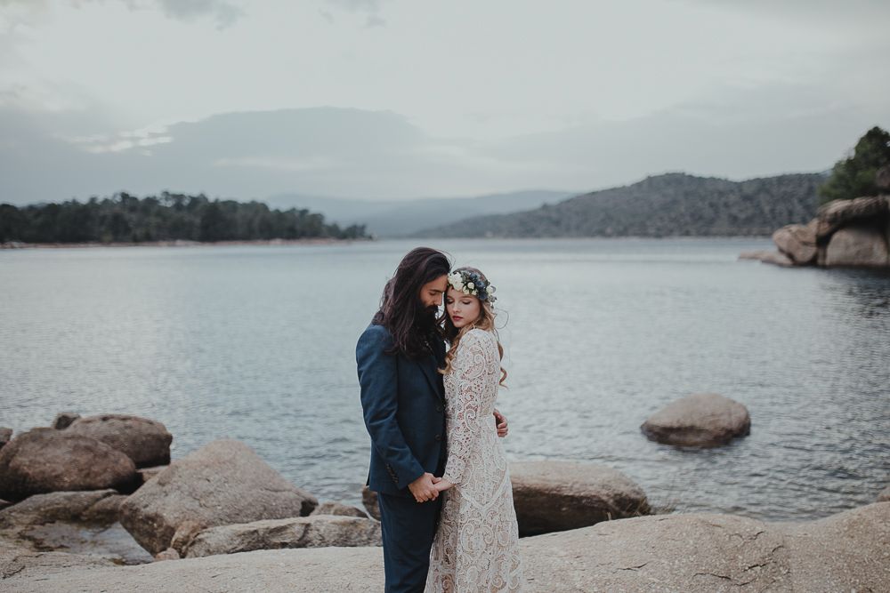 Two people embrace while standing on large rocks by a tranquil mountain lake.