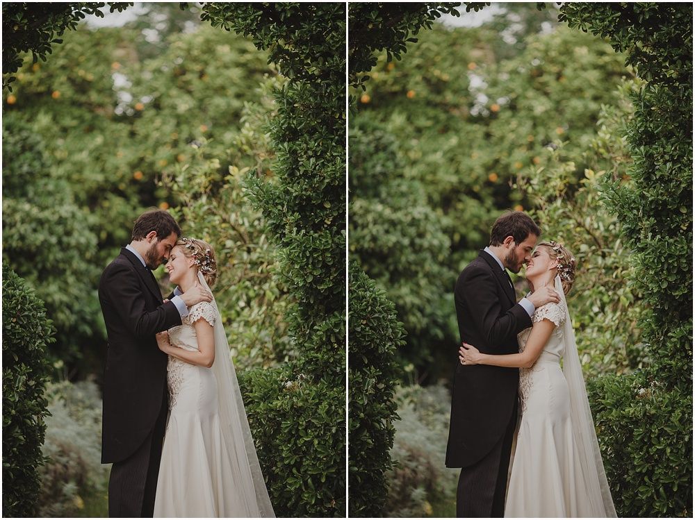 A romantic wedding couple embraces in a garden surrounded by green foliage.