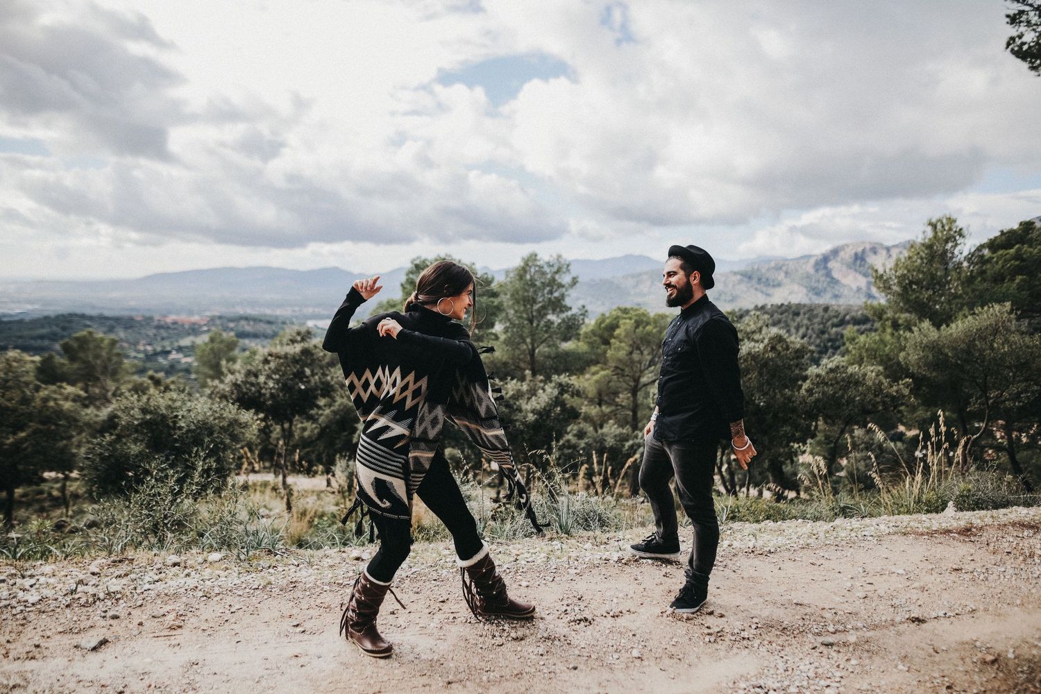 Two people playfully dancing on a mountaintop path with scenic views.