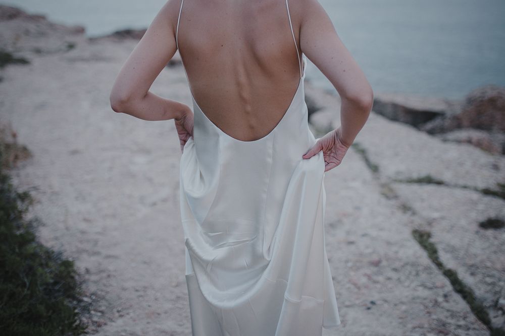 Flowing white backless dress captured from behind on rocky coastal path.