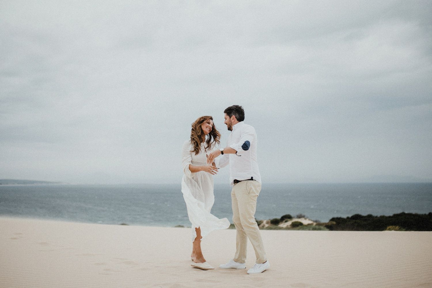 A romantic couple embraces while standing on a sandy beach with an overcast sky and ocean in the background.