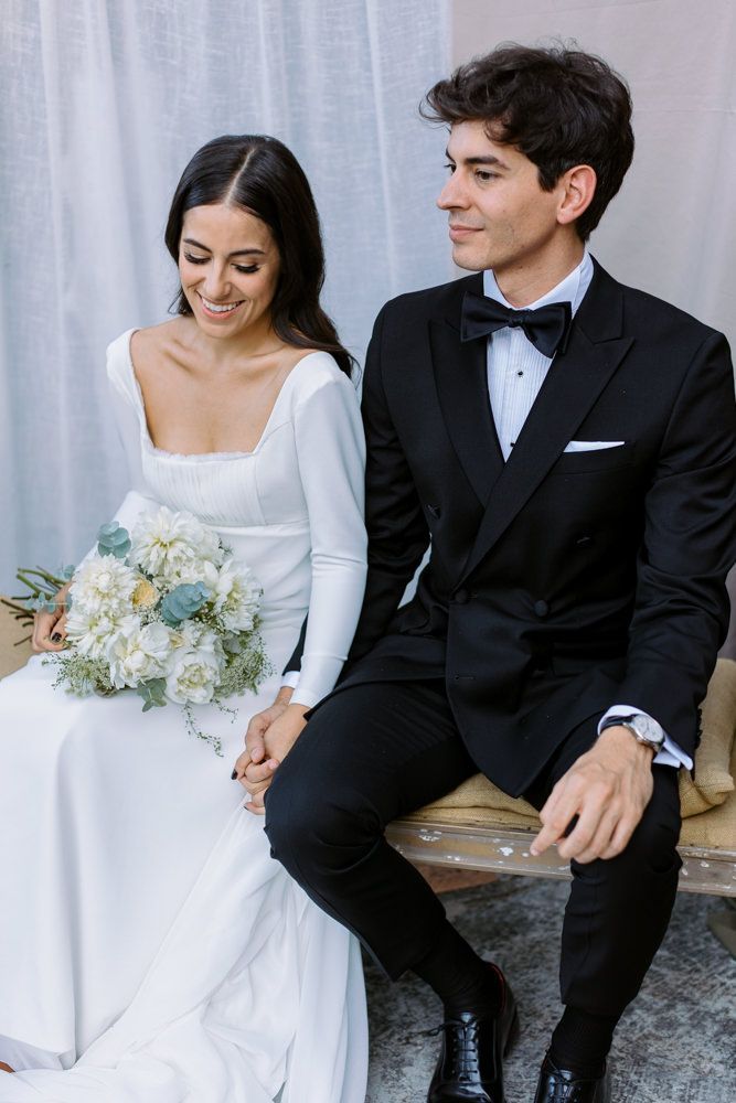 A wedding couple in formal attire sitting together with a bouquet of light blue flowers.