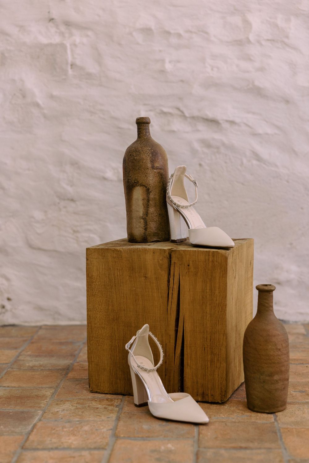 White bridal heels arranged with vintage brown bottles on a wooden block against a white wall.