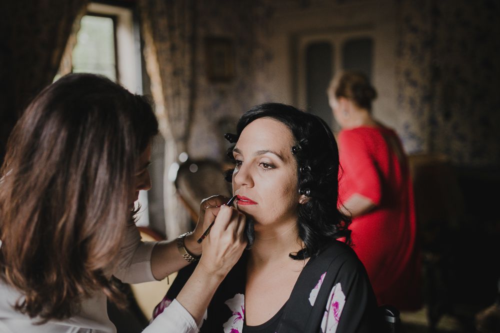 Makeup artist applies cosmetics during wedding preparation in a vintage-styled room.