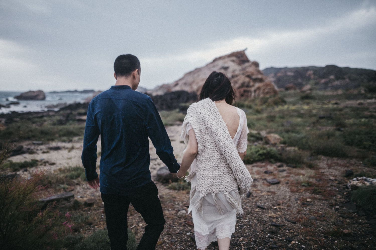 A couple walks hand in hand along a rocky coastal path wearing formal attire.