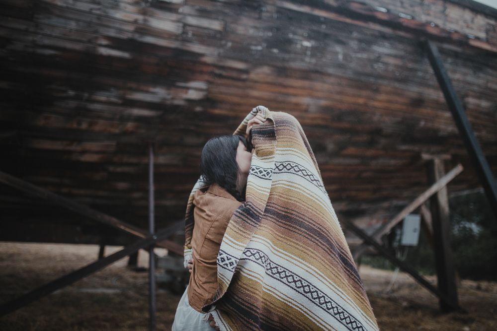 A couple wrapped in a patterned blanket embraces near a wooden structure.