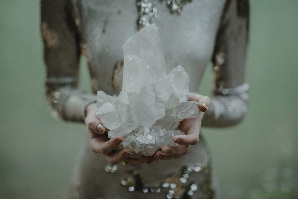 Hands holding large clear quartz crystal clusters against a dreamy green background.
