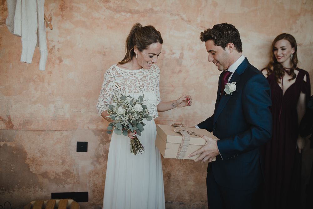 A bride in lace gown and groom in navy suit share a joyful moment against a rustic wall.