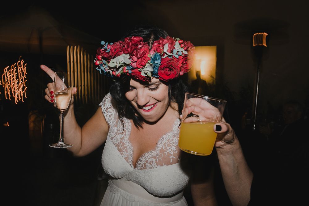 A bride in a white dress and red flower crown holding up a yellow drink at a celebration.
