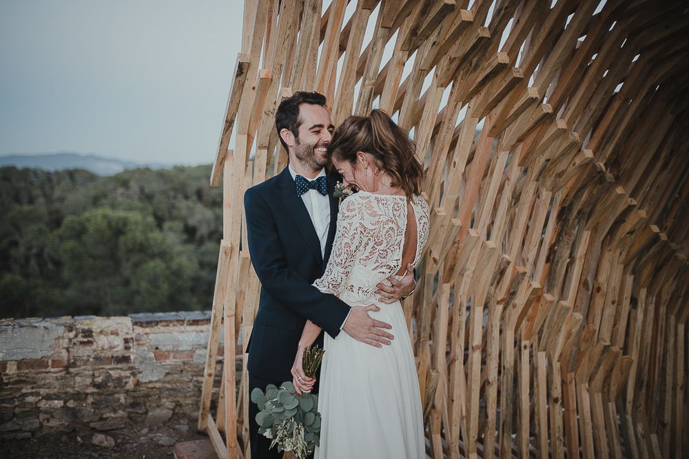 A romantic wedding couple poses against a curved wooden architectural element in a rustic setting.