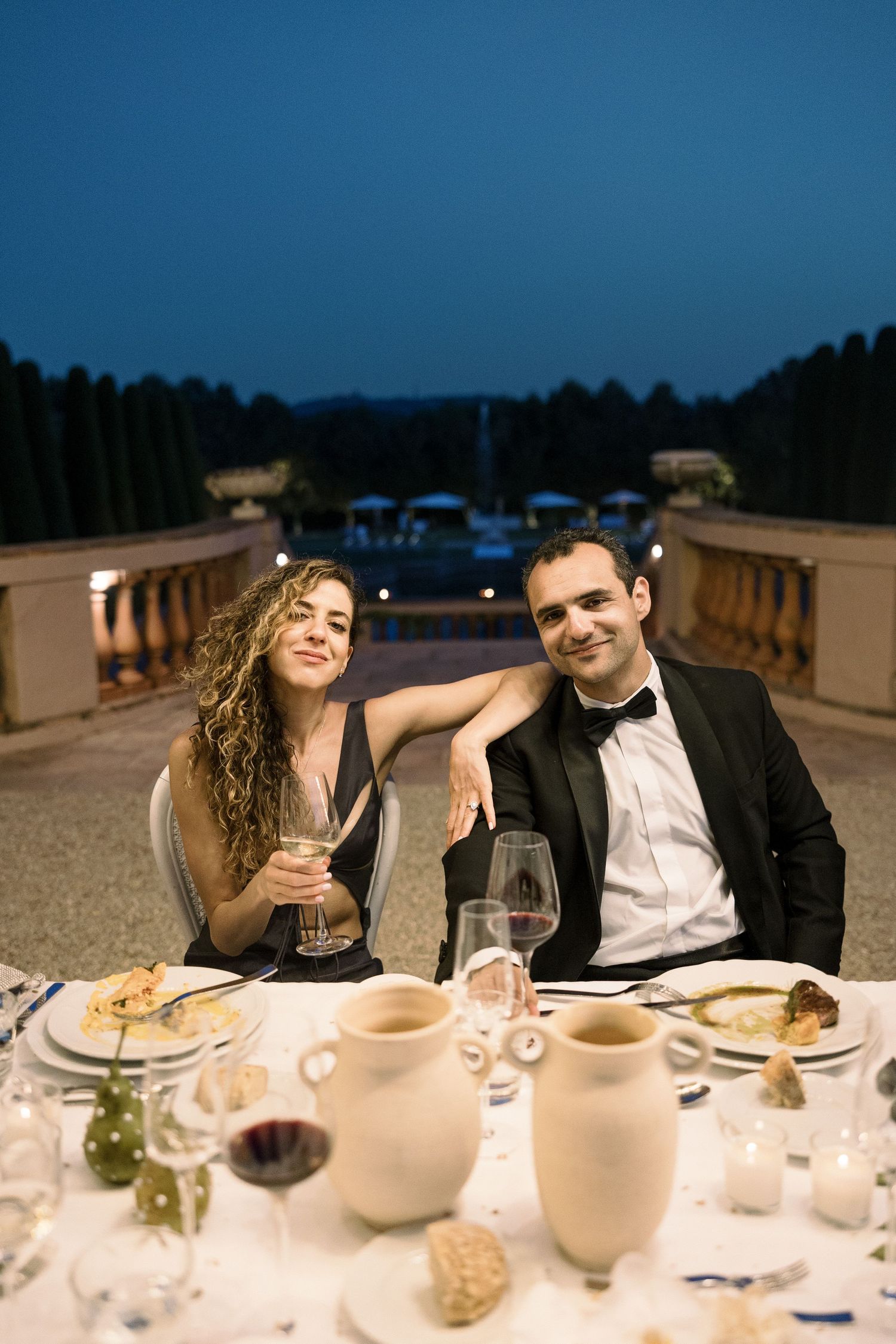Evening wedding reception dinner with couple enjoying wine at decorated outdoor table.