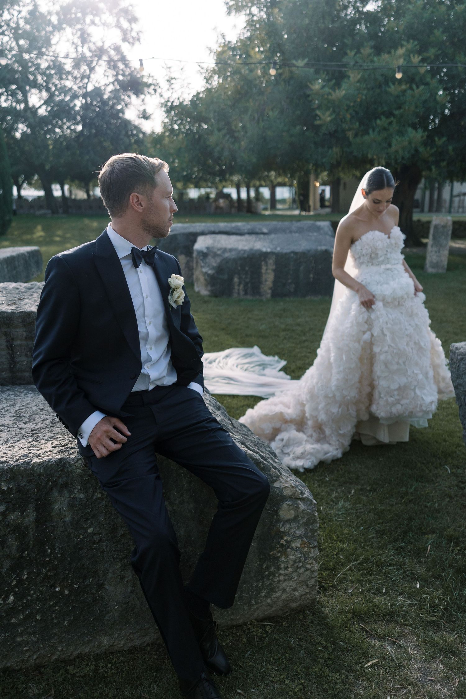 Artistic wedding photo of couple resting on stone wall in garden setting.
