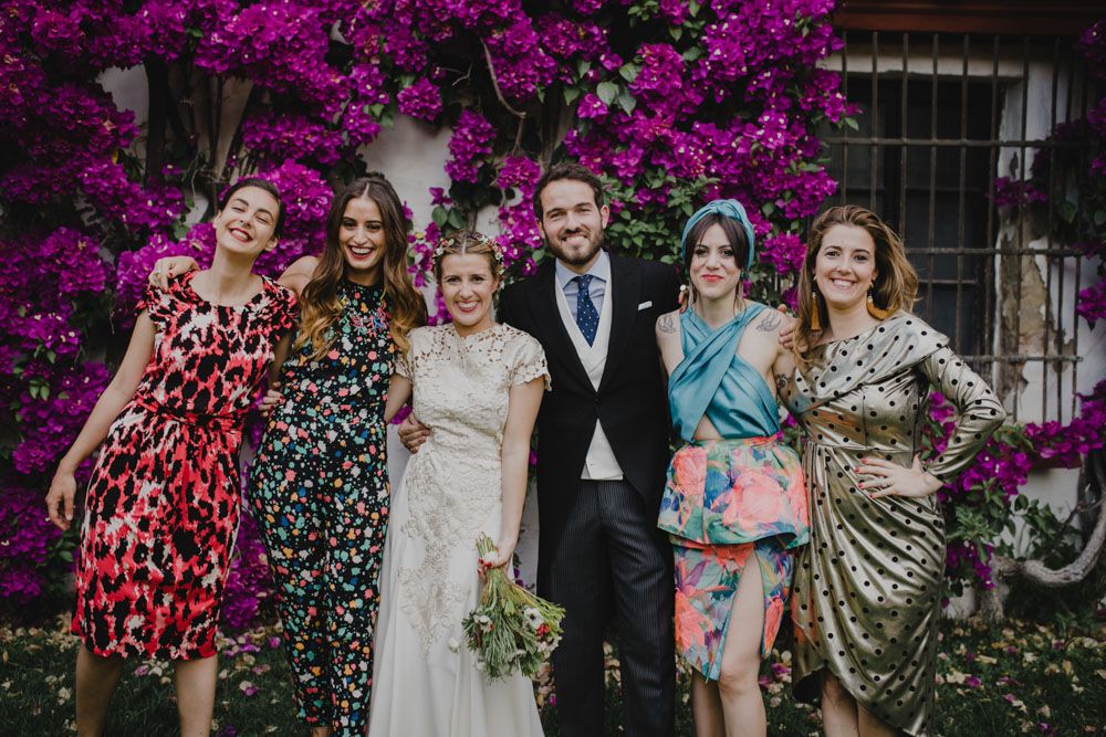 A wedding party poses together against a vibrant purple bougainvillea backdrop wearing colorful dresses.