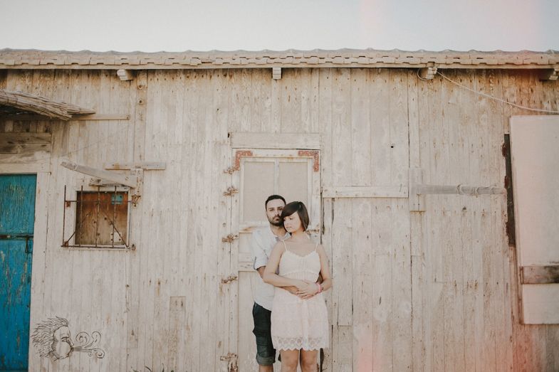A couple embraces in front of a rustic white wooden wall with a blue door.