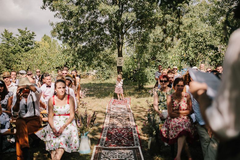 Wedding guests gather on vintage rugs for an outdoor ceremony under green trees.