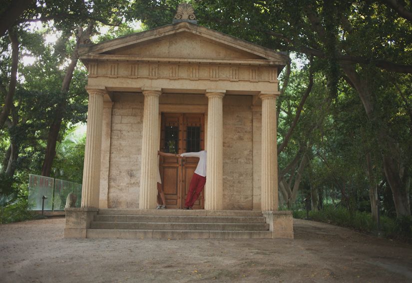Small neoclassical stone temple building with columns surrounded by trees.
