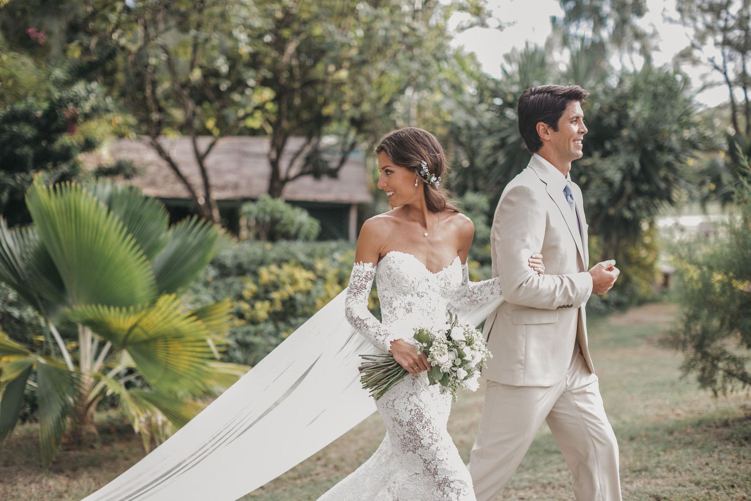 A couple in light-colored wedding attire walks through a tropical garden setting.