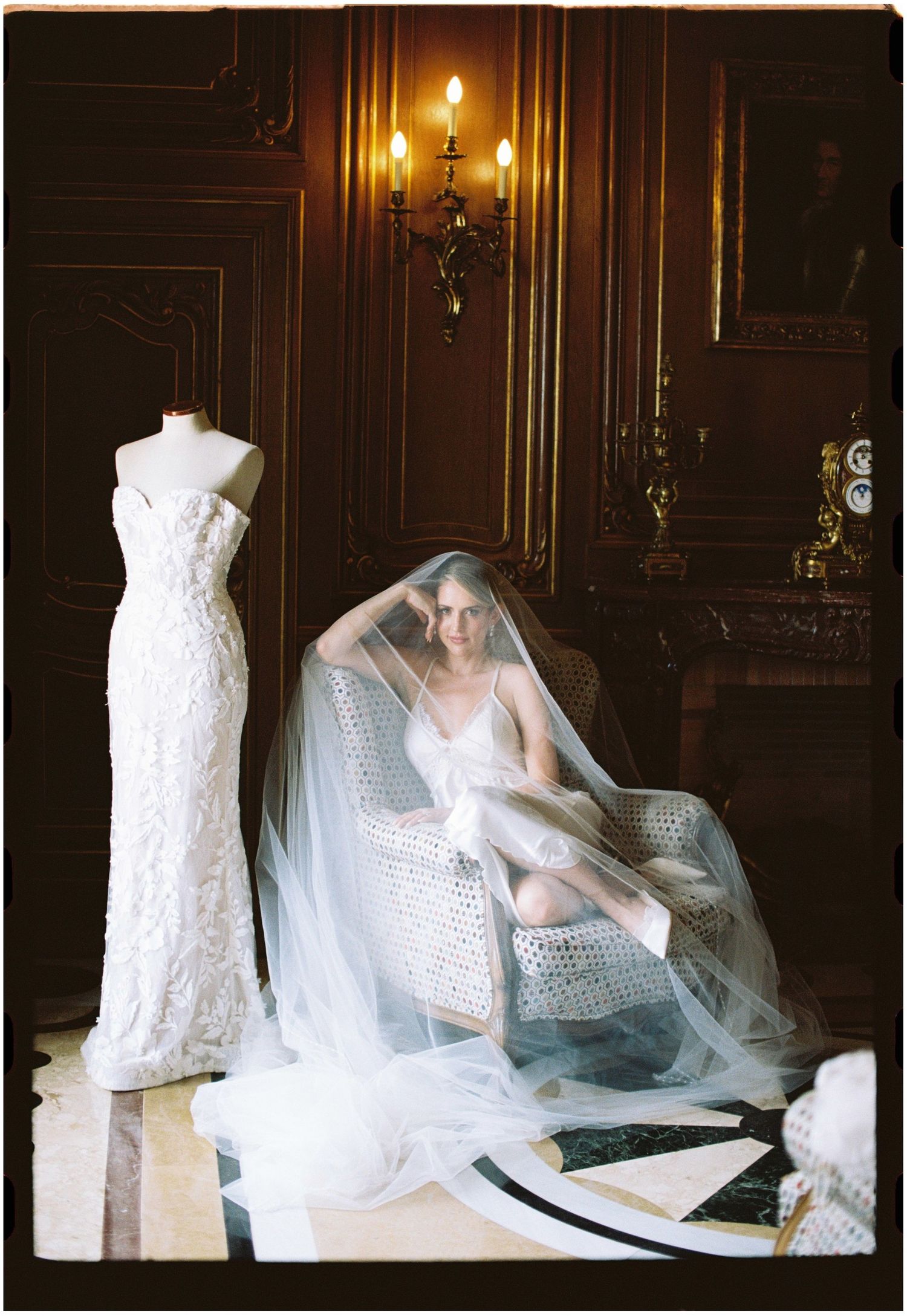 Bride in veil sits in ornate chair beside wedding gown on mannequin in elegant candlelit room.