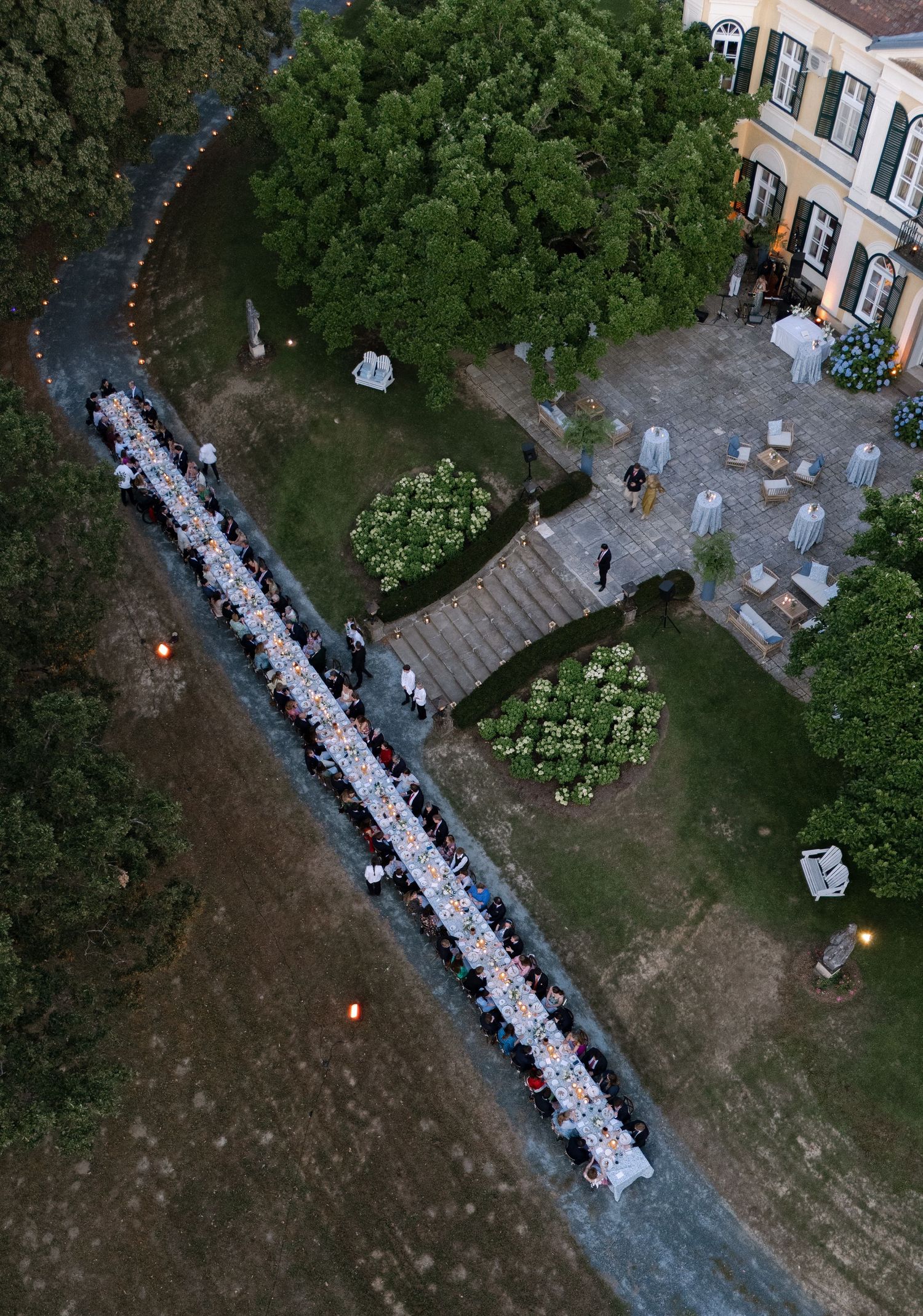 Aerial view of an elegant outdoor wedding reception with long dining tables set in a lush garden beside a grand villa.
