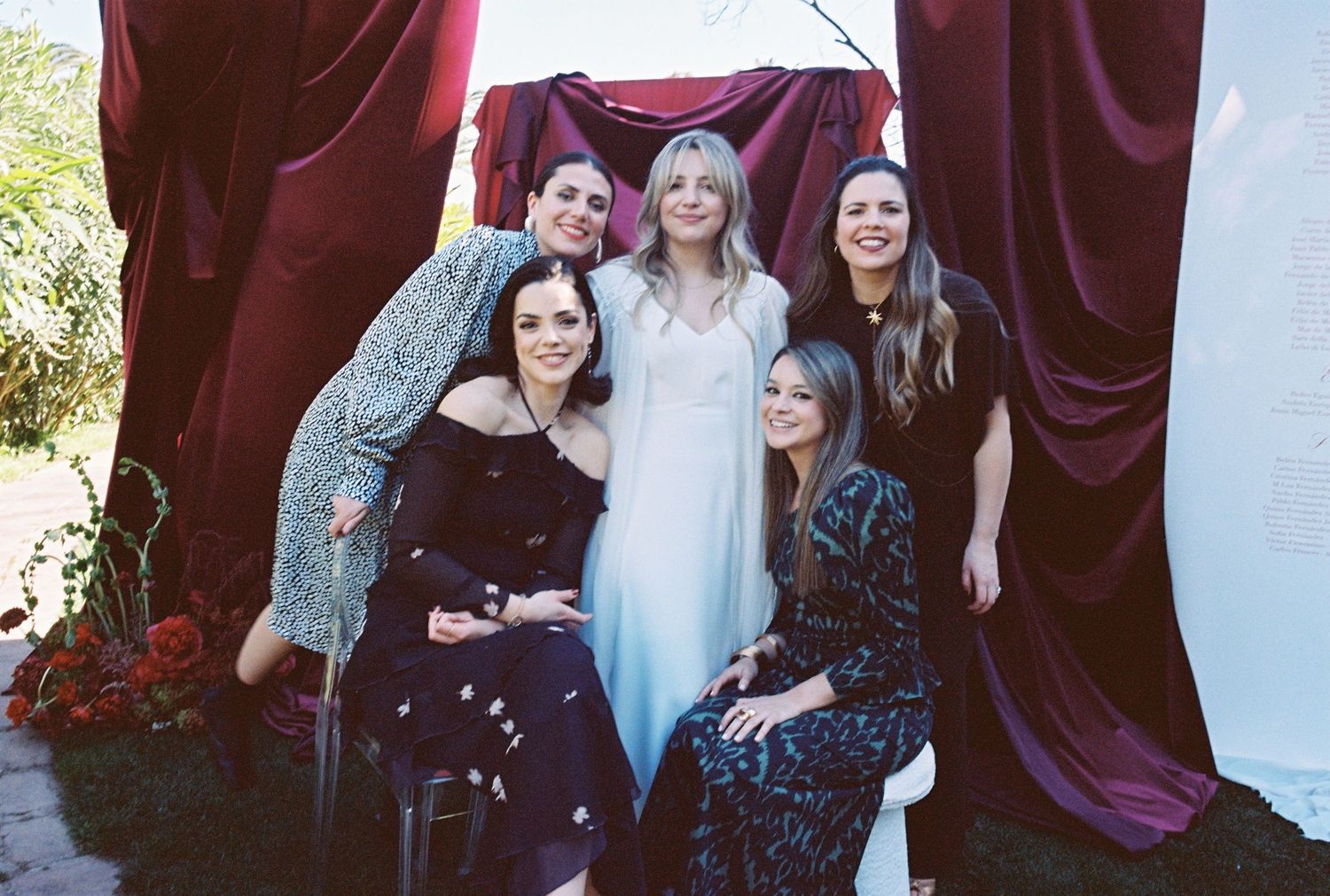 A group poses together against burgundy curtains outdoors wearing dark and light colored dresses.