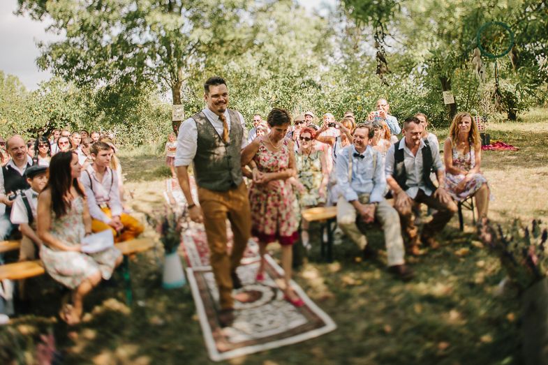 Bohemian outdoor wedding ceremony with guests watching from rustic wooden seating.