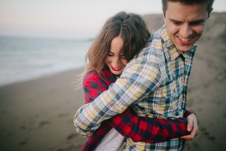 A couple laughs together on a beach, with one wearing a red plaid shirt.