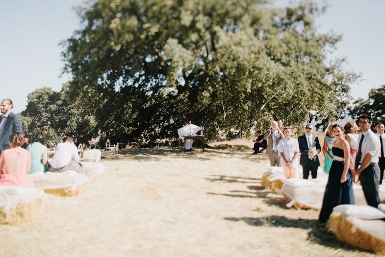 Outdoor wedding ceremony setup under trees with guests seated on hay bales.