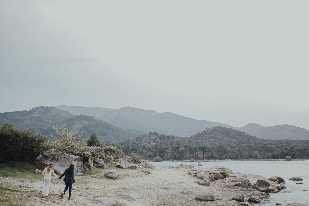 Distant figure walks along rugged shoreline with mountains in background.