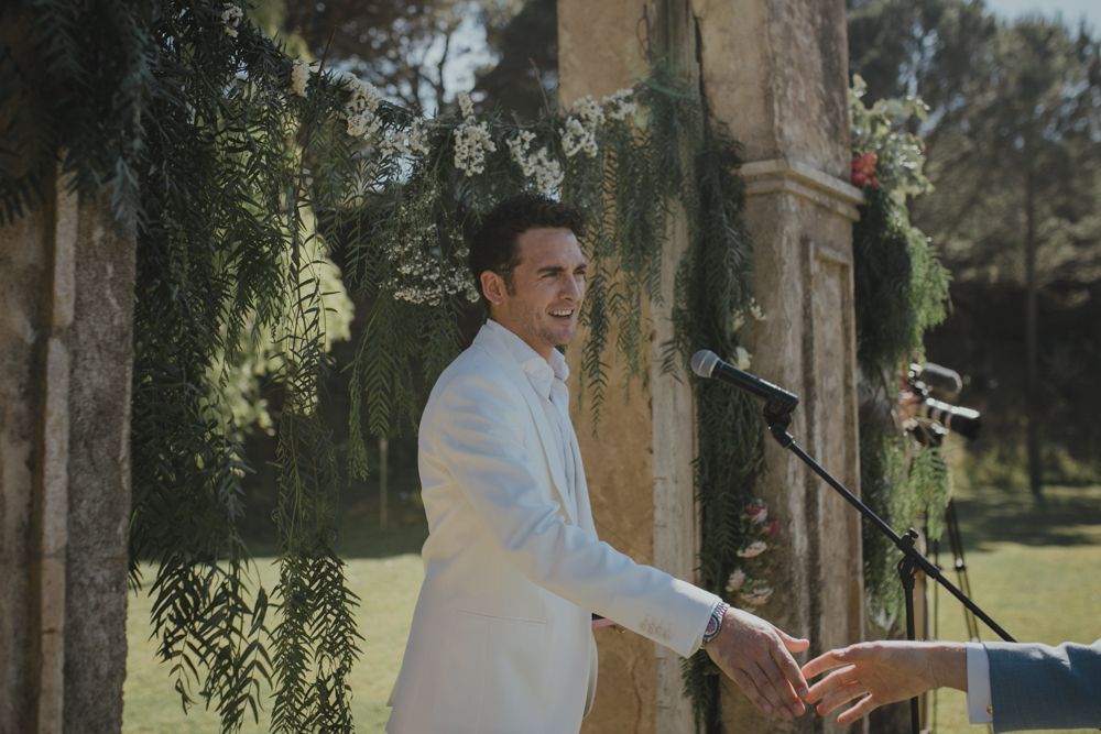 A speaker in a white suit jacket delivering remarks at an outdoor wedding ceremony.