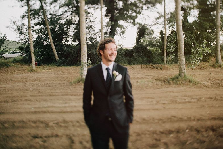 Groom in black suit standing among trees at outdoor wedding venue.