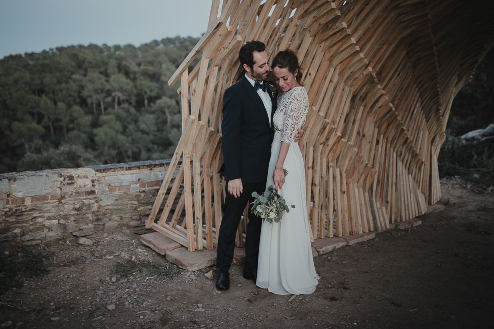 A romantic wedding portrait against a curved wooden structure in a mountainous setting at dusk.