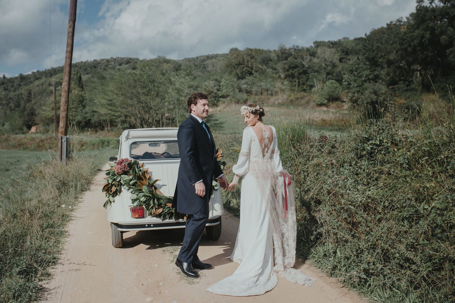 A couple stands next to a vintage pickup truck decorated with floral arrangements on a dirt road in the countryside.