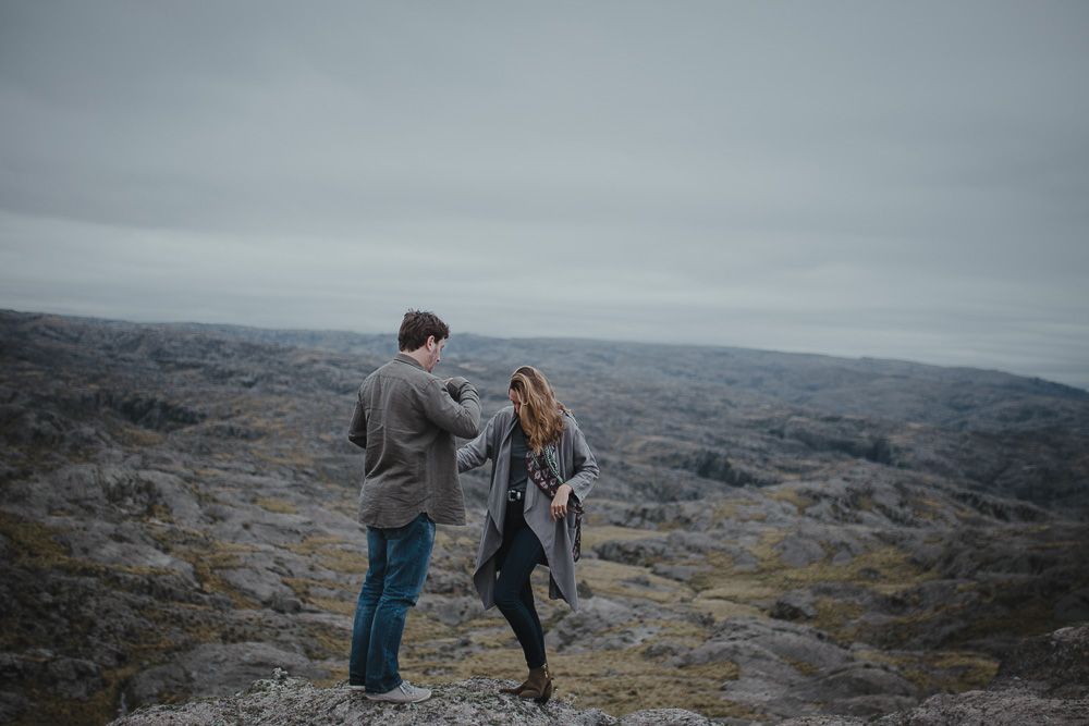 Two people share a dance-like moment on rocky terrain under an overcast sky.