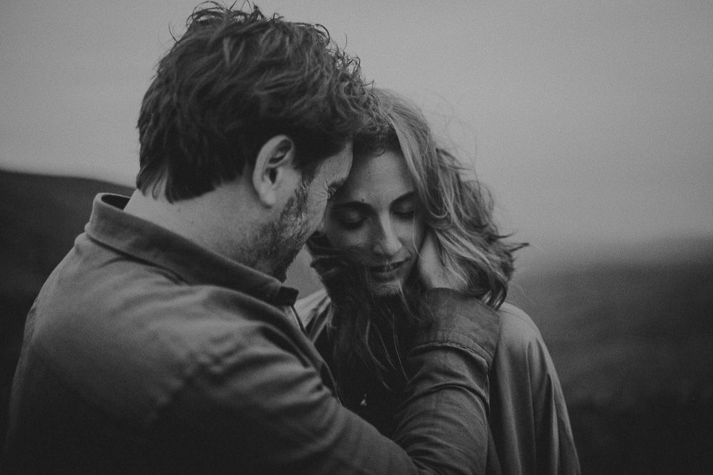 A romantic couple embraces against misty mountain scenery during overcast day.