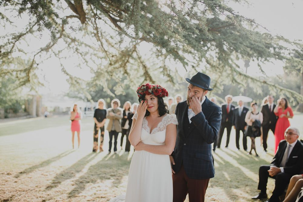 A joyful wedding celebration with guests watching the ceremony under towering trees.