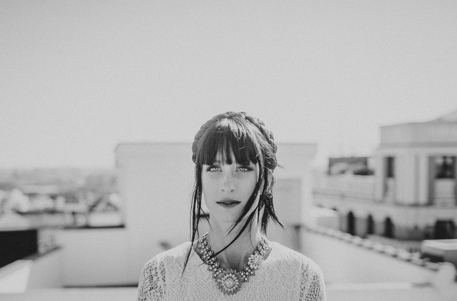 Portrait of person in white dress with dark bangs and tattoos against urban backdrop.