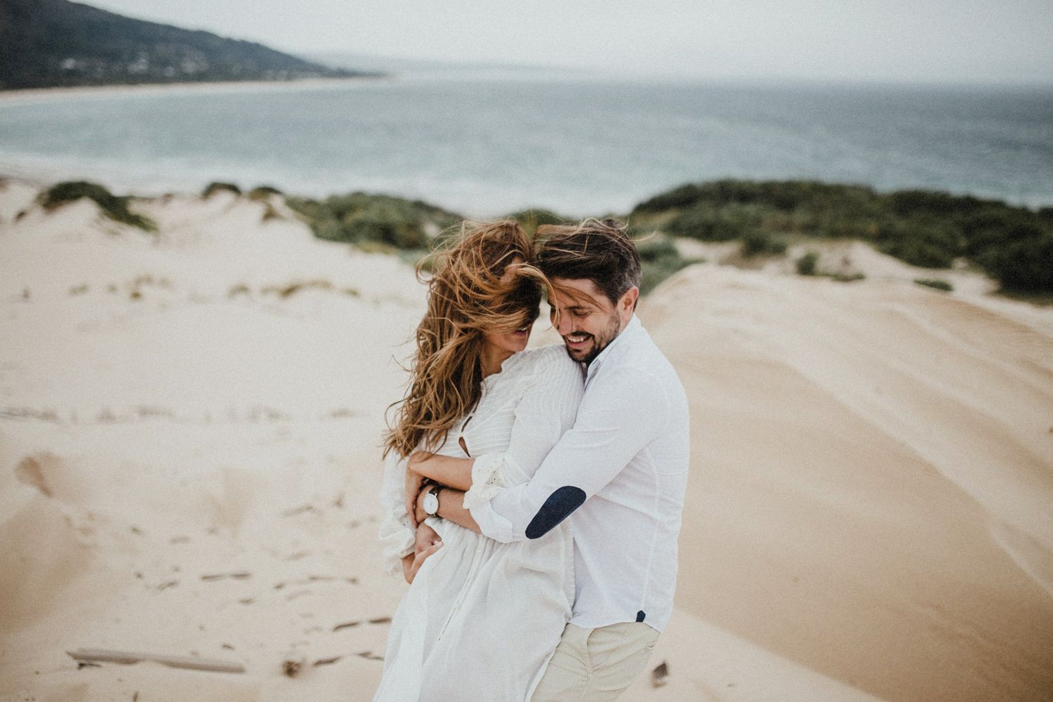 Couple in white embracing on coastal sand dunes with ocean view.