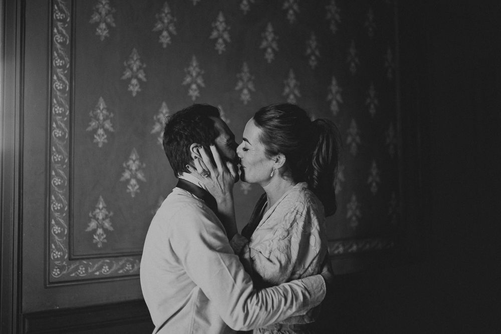 Intimate black and white portrait against patterned wallpaper in dim lighting.