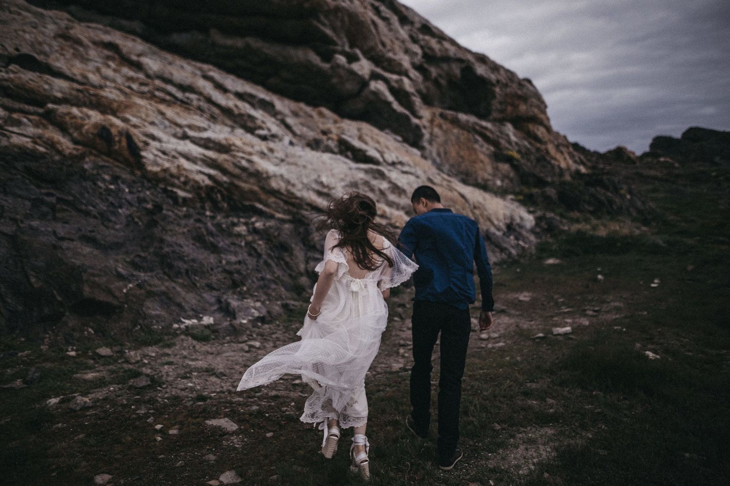 A couple walking hand in hand along a rocky coastal path in wedding attire.